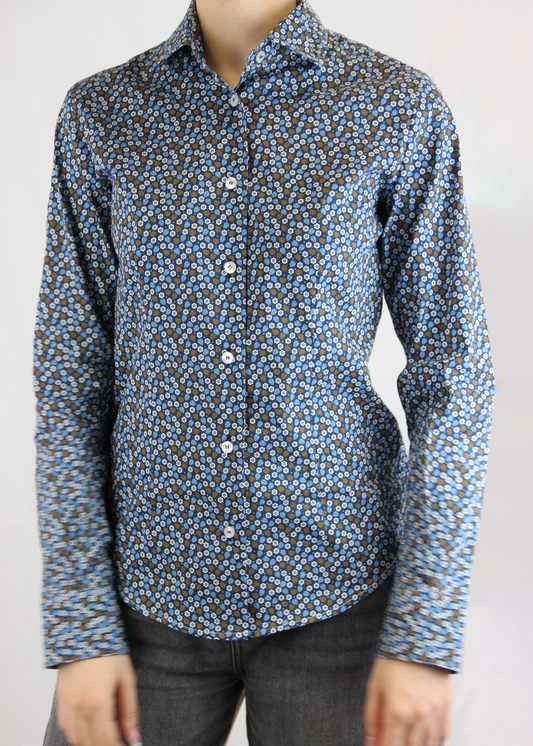 Camicia da donna in cotone con fantasia floreale su sfondo blu. Il design elegante presenta un pattern delicato di piccoli fiori e foglie nei toni dell’azzurro e del marrone, ideale per un look raffinato e senza tempo. Realizzata artigianalmente dalla maglieria Strike Inside Abbigliamento a Genova, questa camicia unisce qualità sartoriale e comfort, perfetta per ogni occasione.