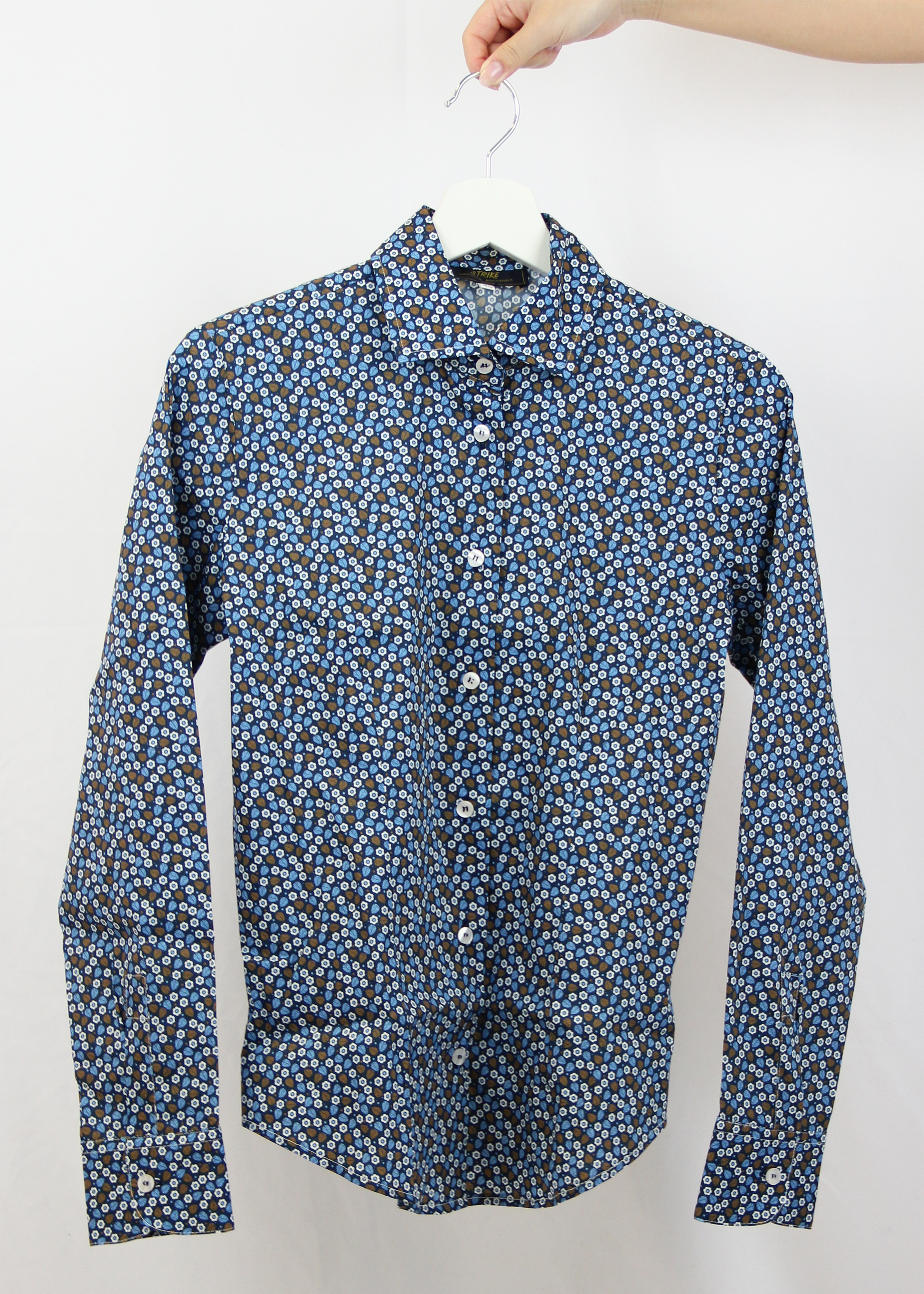 Camicia da donna in cotone con fantasia floreale su sfondo blu. Il design elegante presenta un pattern delicato di piccoli fiori e foglie nei toni dell’azzurro e del marrone, ideale per un look raffinato e senza tempo. Realizzata artigianalmente dalla maglieria Strike Inside Abbigliamento a Genova, questa camicia unisce qualità sartoriale e comfort, perfetta per ogni occasione.
