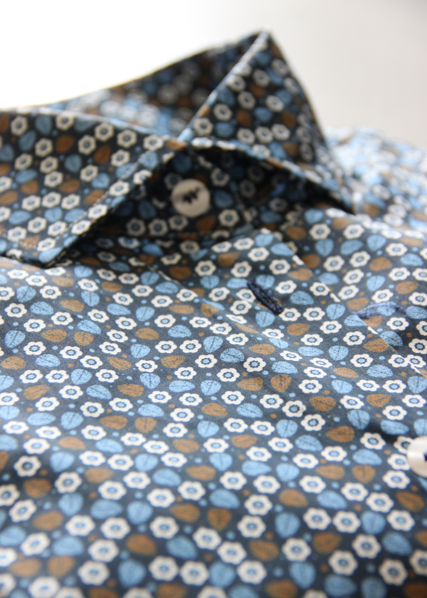 Camicia da donna in cotone con fantasia floreale su sfondo blu. Il design elegante presenta un pattern delicato di piccoli fiori e foglie nei toni dell’azzurro e del marrone, ideale per un look raffinato e senza tempo. Realizzata artigianalmente dalla maglieria Strike Inside Abbigliamento a Genova, questa camicia unisce qualità sartoriale e comfort, perfetta per ogni occasione.