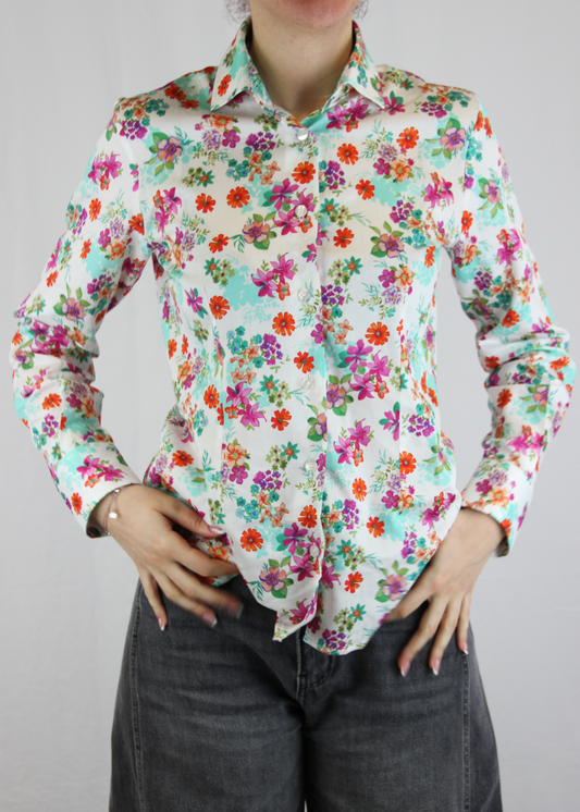 Camicia da donna in cotone dal taglio classico, realizzata artigianalmente da Strike Inside. Il tessuto leggero e traspirante presenta una vivace fantasia floreale su sfondo bianco, decorato con fiori multicolore nei toni del rosso, fucsia, verde e viola. Il design con colletto tradizionale e chiusura con bottoni la rende un capo versatile, perfetto per un look fresco e raffinato. Un'elegante espressione di artigianalità e stile, ideale per aggiungere un tocco di colore al guardaroba.