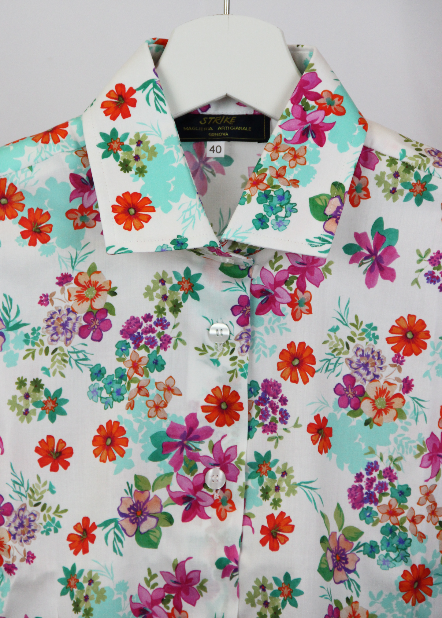 Camicia da donna in cotone dal taglio classico, realizzata artigianalmente da Strike Inside. Il tessuto leggero e traspirante presenta una vivace fantasia floreale su sfondo bianco, decorato con fiori multicolore nei toni del rosso, fucsia, verde e viola. Il design con colletto tradizionale e chiusura con bottoni la rende un capo versatile, perfetto per un look fresco e raffinato. Un'elegante espressione di artigianalità e stile, ideale per aggiungere un tocco di colore al guardaroba.