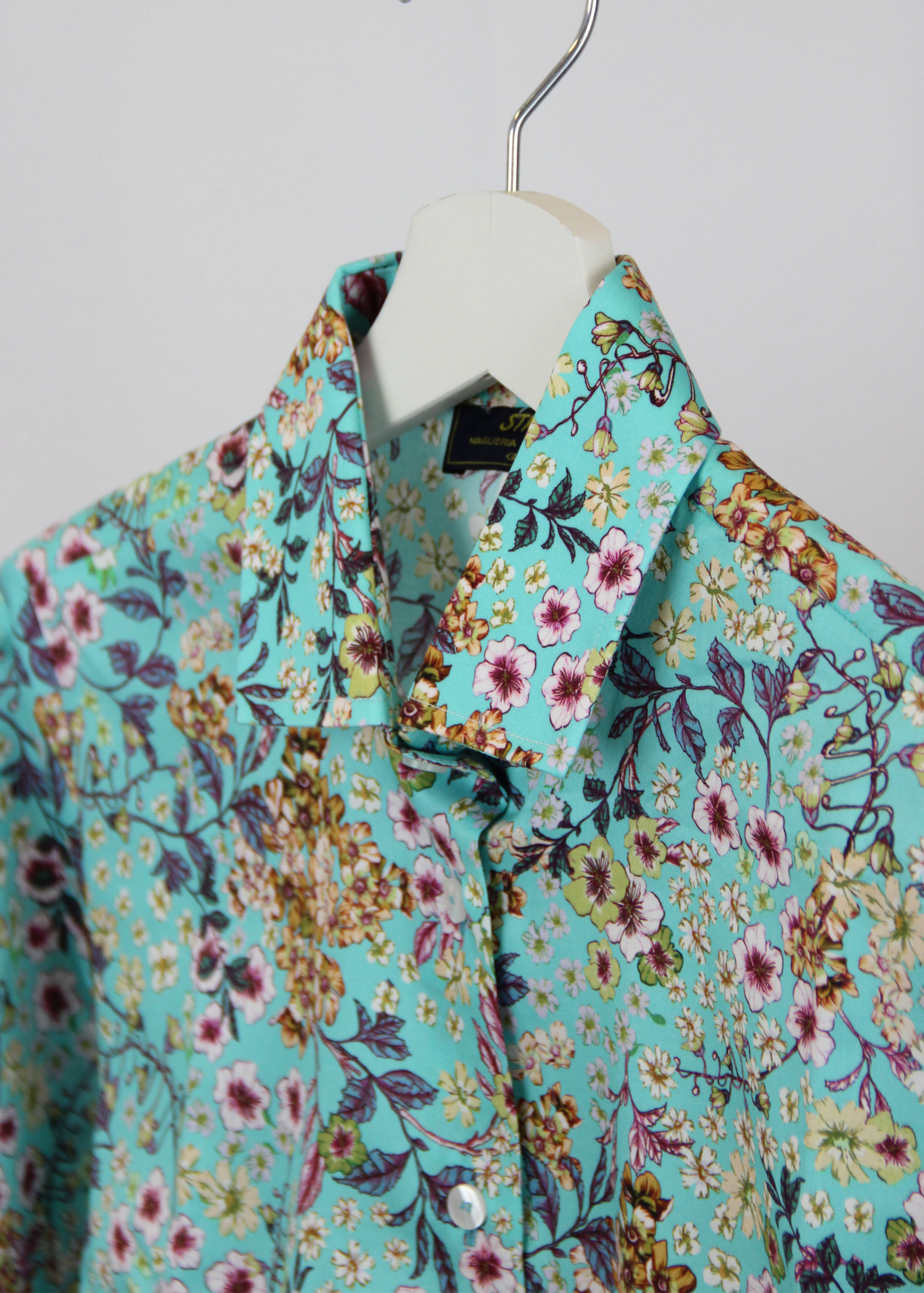 Camicia da donna in cotone dal taglio classico, realizzata artigianalmente da Strike Inside. Il tessuto leggero e traspirante presenta una delicata fantasia floreale con uno sfondo azzurro-verde acqua, arricchito da fiori rosa, gialli e viola. Il design con colletto tradizionale e chiusura con bottoni la rende perfetta per un look fresco e sofisticato. Un capo unico che unisce artigianalità e stile, ideale per donare un tocco di colore e femminilità al guardaroba.